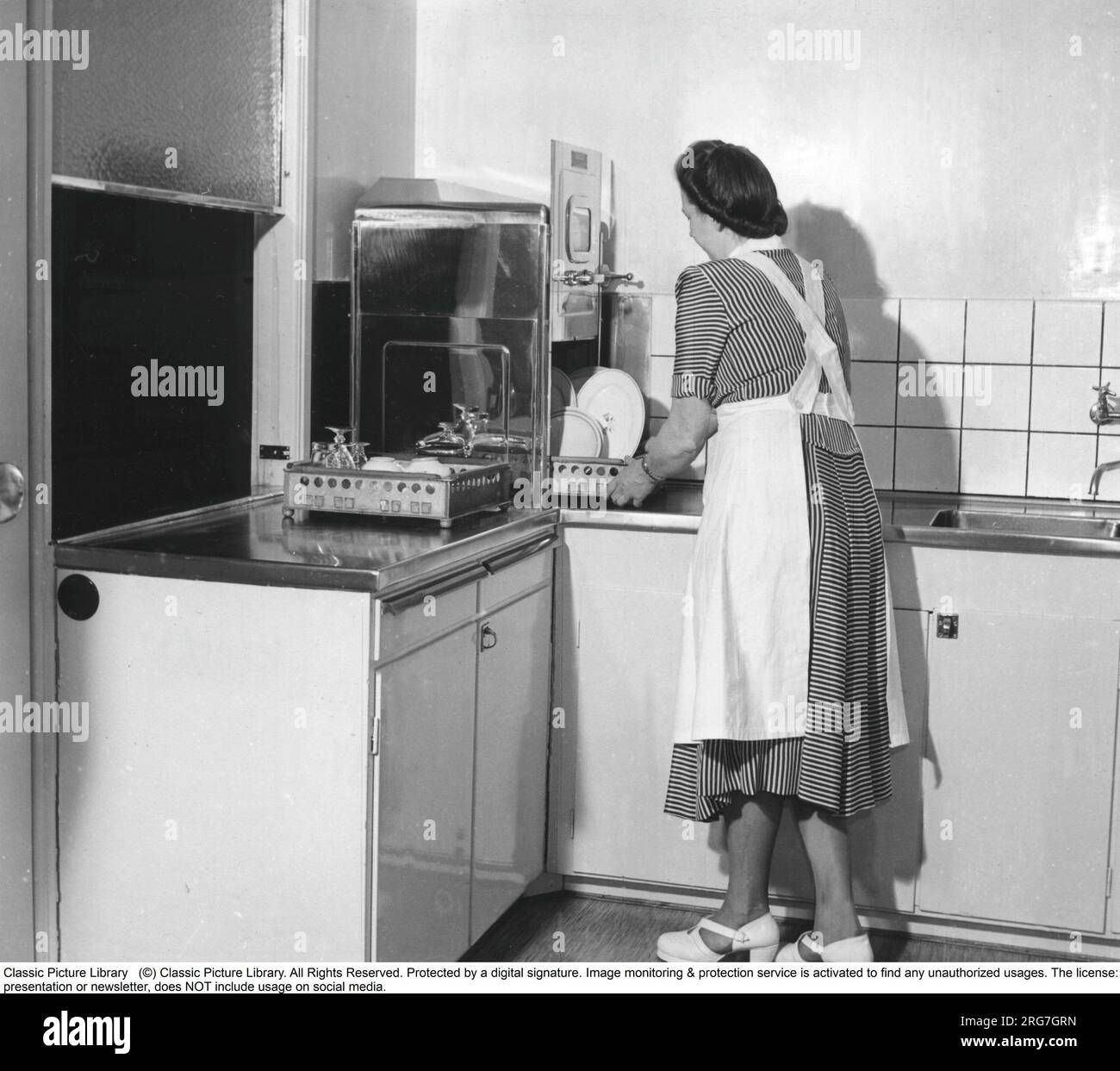 Küche in den 1950er. Eine Frau wird in der 1950er-Küche gesehen, die die Spülmaschine benutzt und auf dem Waschbecken steht. Ein zeittypisches Modell mit Edelstahlabdeckung. Schweden 1952. Stockfoto