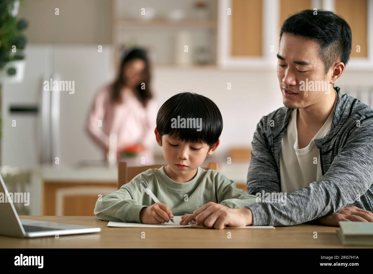 Junger asiatischer Vater, der an einem Tisch sitzt und den Sohn zu Hause unterrichtet, während die Mutter in der Küche kocht Stockfoto