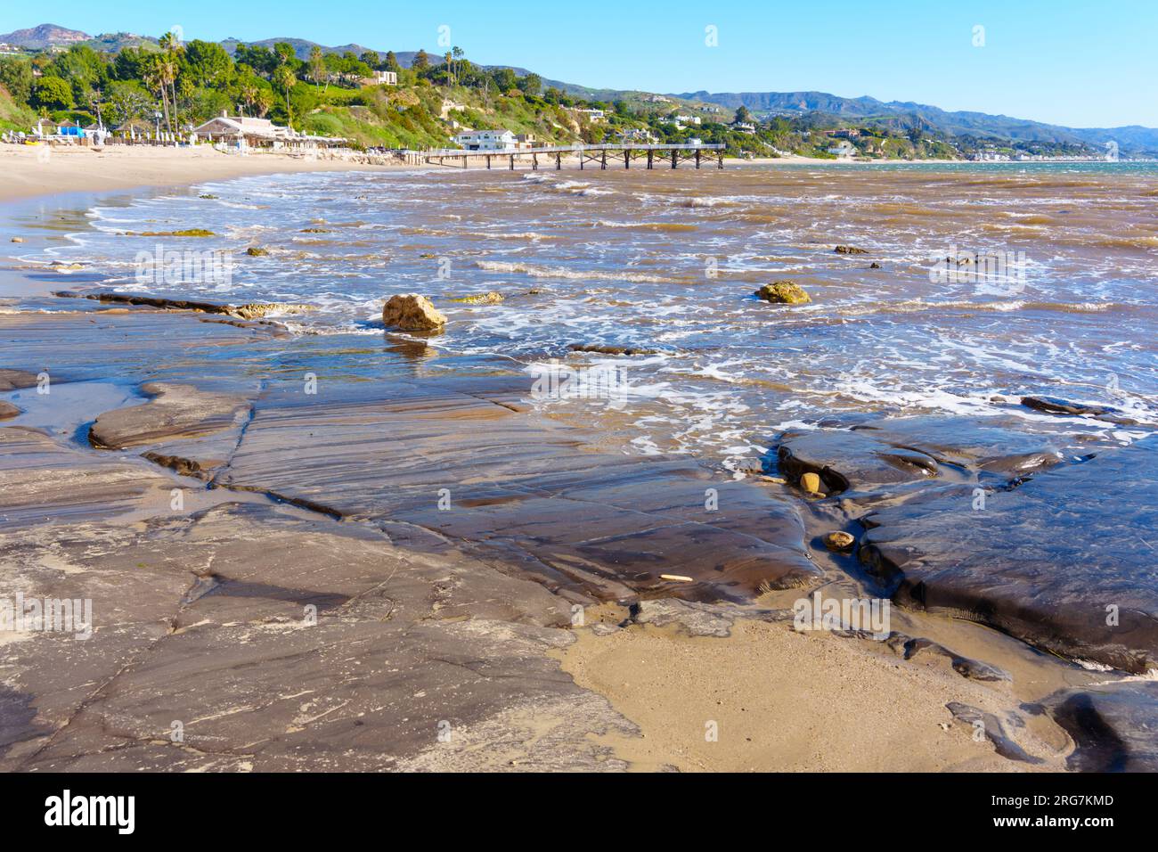 Die ungezähmte Pracht des Malibu Beach zeigt seine rohe und natürliche Schönheit mit felsiger Küste, hölzernem Pier, grünen Hügeln und blauem Himmel. Stockfoto