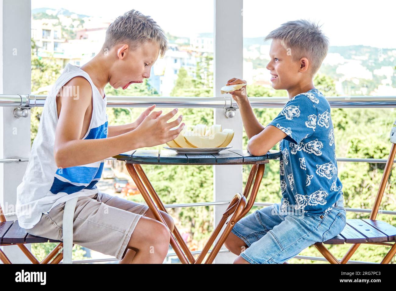 Kinder und Jugendliche genießen frische Melonen auf der Hotelterrasse. Geschwister essen Melonenscheiben mit aufgeregtem Ausdruck Stockfoto