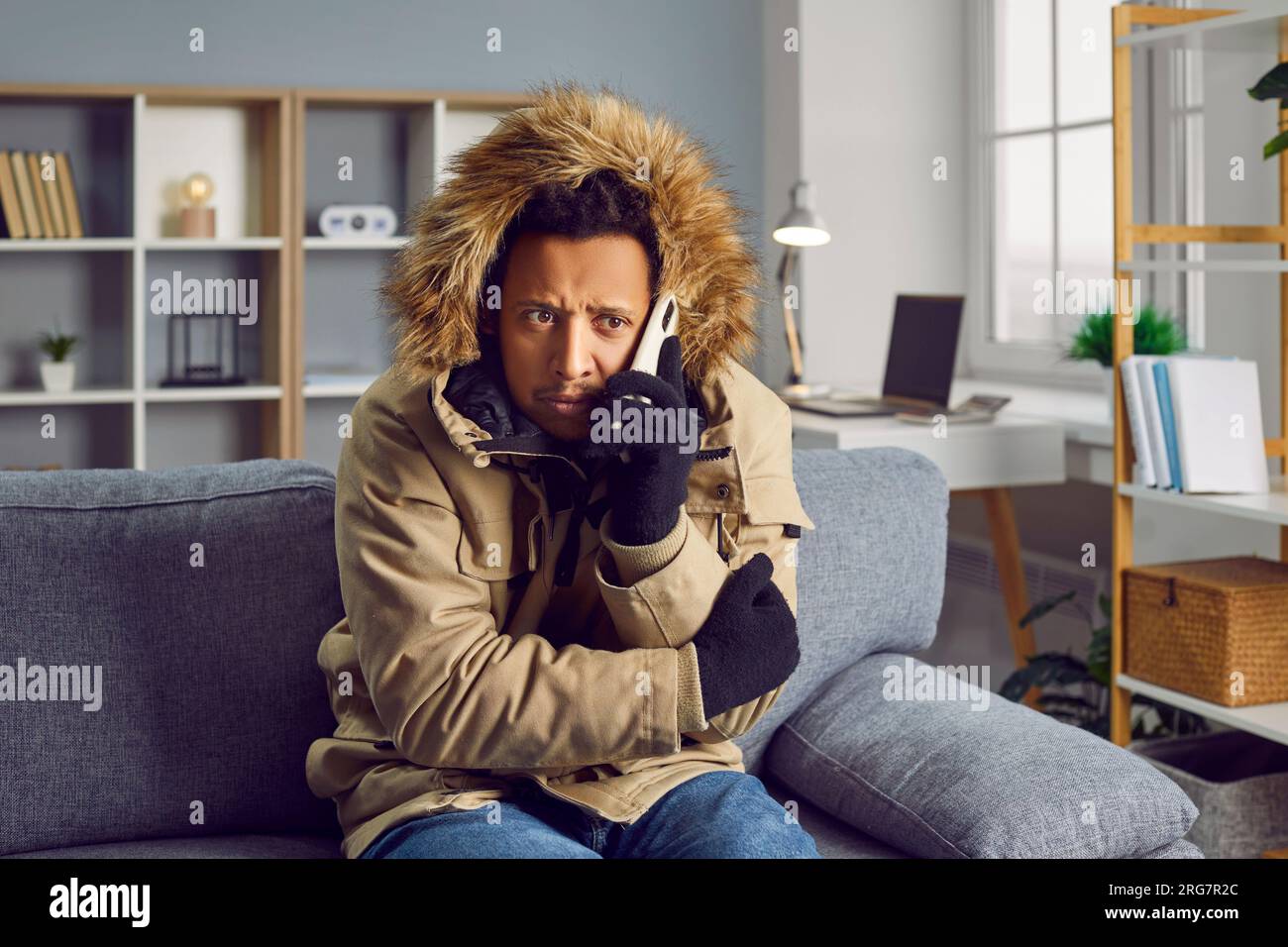 Junger gefrorener Mann, der zu Hause in einem Wintermantel auf dem Sofa sitzt und telefoniert. Stockfoto