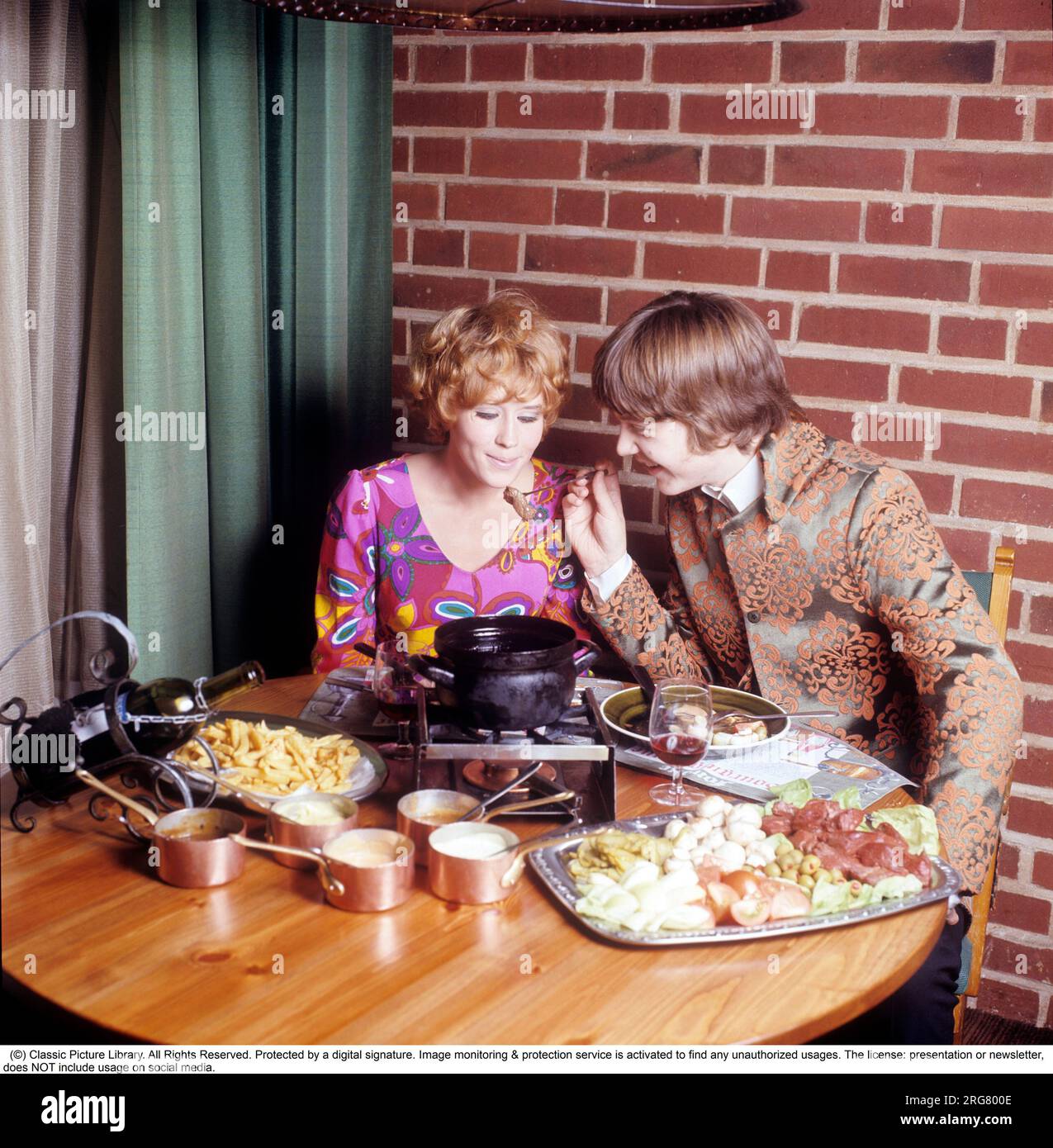 Fondue. Ein Gericht, das seinen Durchbruch in den 60s und 70s Jahren hatte. Ein Fondue-Topf mit heißem Öl steht in der Mitte des Tisches und man taucht Fleisch hinein, um sich selbst zu kochen und zu essen. Sie trägt eine sehr farbenfrohe und gemusterte Langarmbluse, die typisch für die späten 60er und 70er Jahre war. Er trug eine Pop-inspirierte Jacke, die sich nicht von der Kleidung unterscheidet, die die Sänger damals trugen. Schweden 1970. Conard Ref CV49-5 Stockfoto