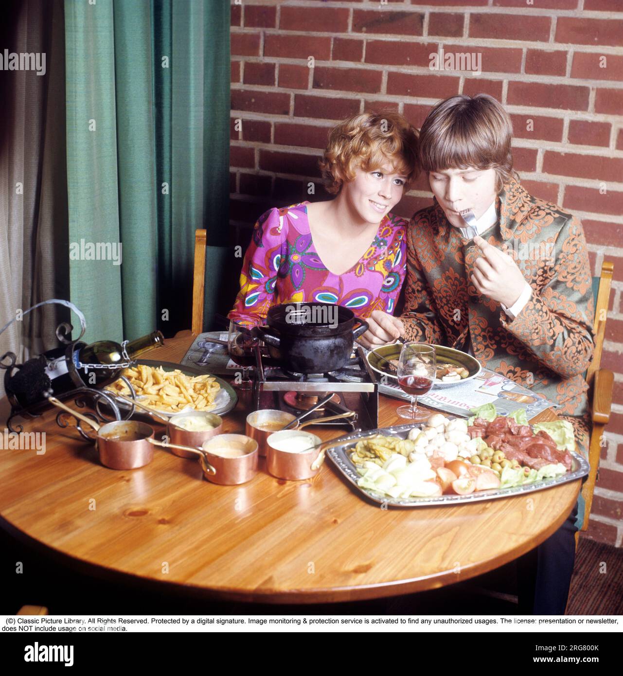 Fondue. Ein Gericht, das seinen Durchbruch in den 60s und 70s Jahren hatte. Ein Fondue-Topf mit heißem Öl steht in der Mitte des Tisches und man taucht Fleisch hinein, um sich selbst zu kochen und zu essen. Sie trägt eine sehr farbenfrohe und gemusterte Langarmbluse, die typisch für die späten 60er und 70er Jahre war. Er trug eine Pop-inspirierte Jacke, die sich nicht von der Kleidung unterscheidet, die die Sänger damals trugen. Schweden 1970. Conard Ref CV49-5 Stockfoto