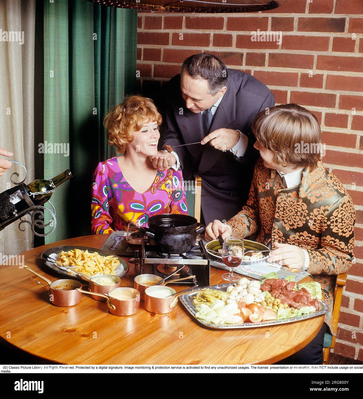 Fondue. Ein Gericht, das seinen Durchbruch in den 60s und 70s Jahren hatte. Ein Fondue-Topf mit heißem Öl steht in der Mitte des Tisches und man taucht Fleisch hinein, um sich selbst zu kochen und zu essen. Sie trägt eine sehr farbenfrohe und gemusterte Langarmbluse, die typisch für die späten 60er und 70er Jahre war. Er trug eine Pop-inspirierte Jacke, die sich nicht von der Kleidung unterscheidet, die die Sänger damals trugen. Schweden 1970. Conard Ref CV49-5 Stockfoto