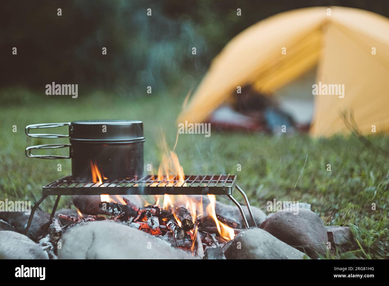 Kessel über brennendem Lagerfeuer mit Zelt auf grünem Wald am warmen Abend. Im Sommer oder Herbst wird am Lagerfeuer gekocht. Konzept der Werbung Stockfoto