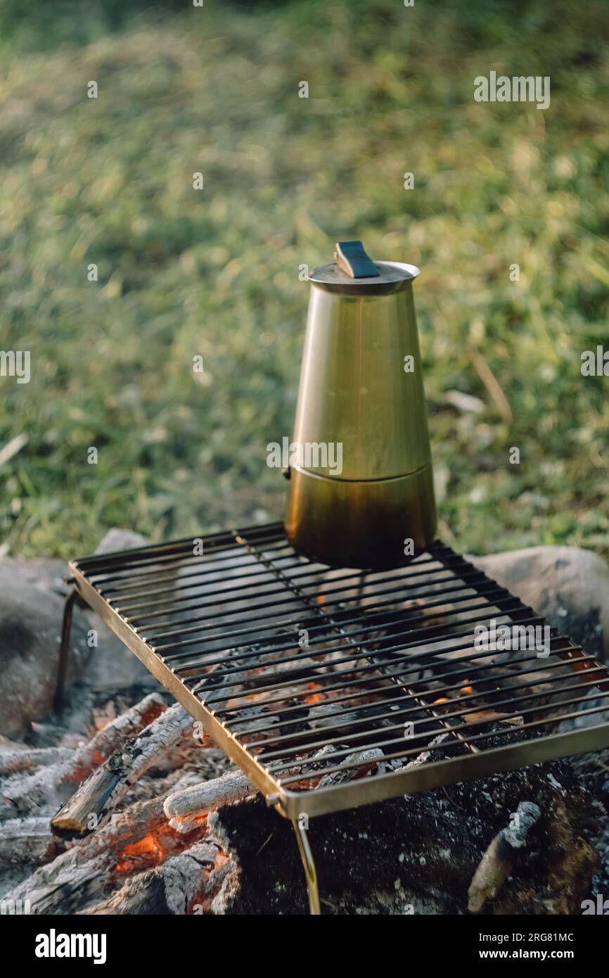 Kaffeezubereitung im Freien am Lagerfeuer im wunderschönen Sonnenaufgang im sommerlichen grünen Park. Campingatmosphäre, Natur- und Outdoor-Lifestyle-Konzept. Lagerfeuer Stockfoto