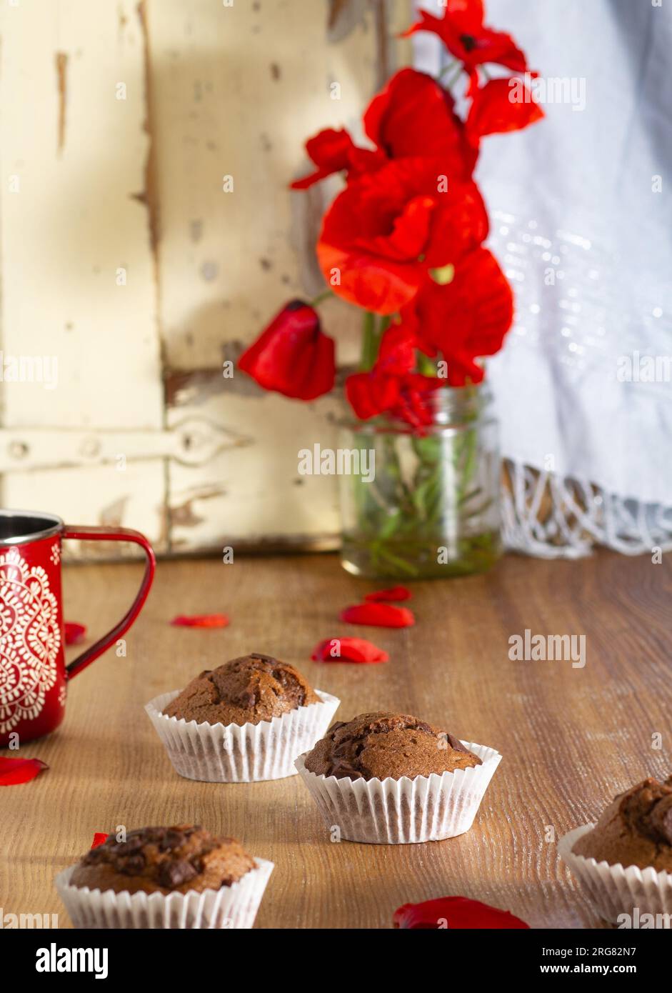 Nahaufnahme des köstlichen Stilllebens von dunklen Schokoladen-Cupcakes, die zu Hause hergestellt werden, umgeben von roten Mohnblüten auf einem rustikalen Holztisch und Details aus Glas Stockfoto