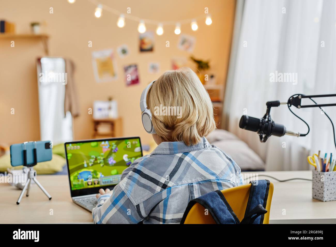Rückansicht eines blonden Schuljungen, der auf einer Laptop-Tastatur tippt, während er in seiner häuslichen Umgebung vor der Kamera am Schreibtisch sitzt und Videospiele spielt Stockfoto