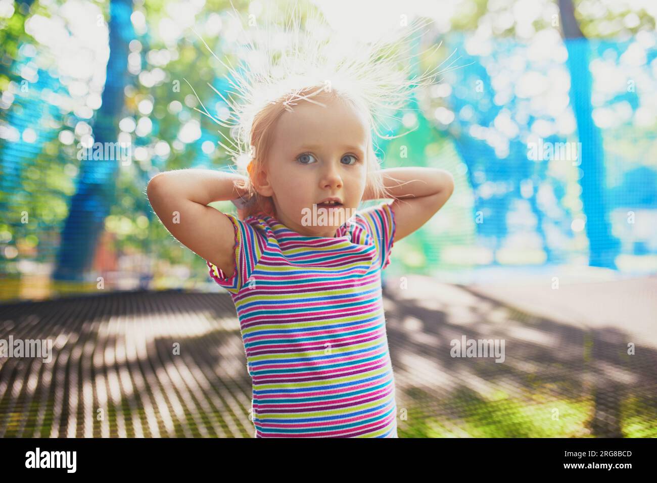 Ein bezauberndes kleines Mädchen, das Spaß im Abenteuerpark hat. Kind mit elektrisiertem Haar auf Baumkronen-Netztrampolin. Aktivitäten im Freien für kleine Kinder Stockfoto