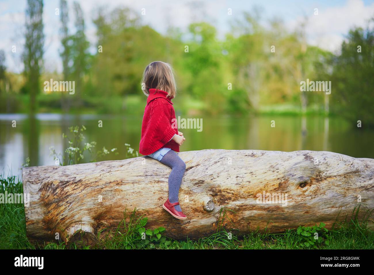 Ein bezauberndes Vorschulmädchen, das auf einem Baumstamm sitzt und Spaß im Frühlingswald oder Park hat. Kind genießt Natur an einem Frühlingstag. Aktivitäten im Freien für Kinder Stockfoto