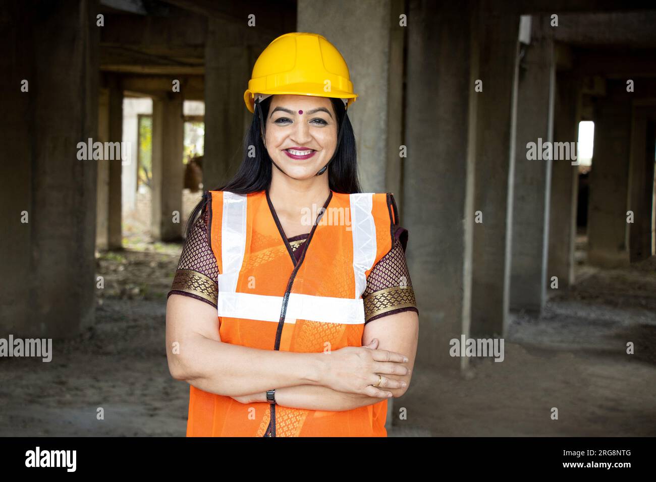 Porträt einer selbstbewussten jungen, wunderschönen indischen Bauingenieurin, die auf der Baustelle einen Helm und eine Weste trägt, die Arme tragend. Stockfoto