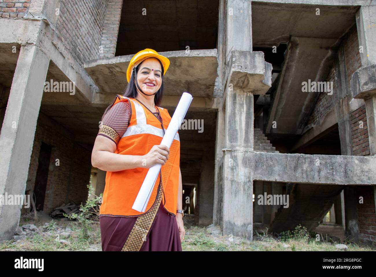 Porträt einer selbstbewussten jungen, wunderschönen indischen Bauingenieurin oder Architektin, die Helm und Weste trägt und einen Papierentwurf auf der Baustelle si hält Stockfoto