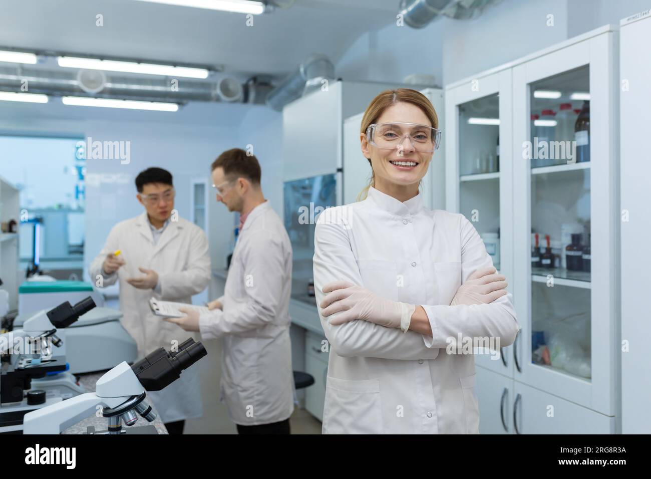 Porträt einer jungen Wissenschaftlerin, Chemikerin, Ärztin, die im Labor der Klinik steht, ihre Arme auf der Brust kreuzt und in die Kamera lächelt. Im Hintergrund, männliche Kollegen. Stockfoto