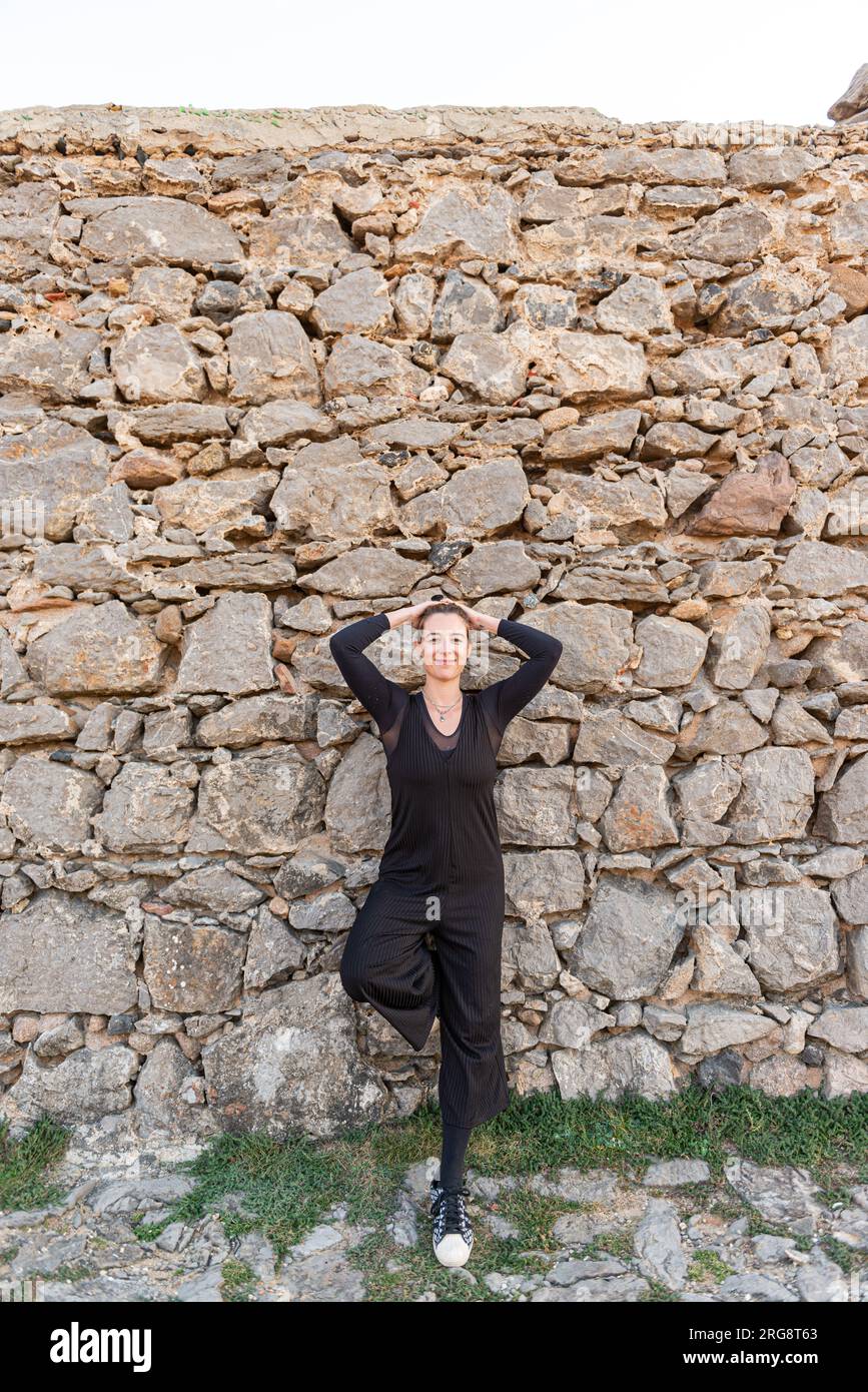 Junge Frau, die legeres schwarzes Outfit trägt und vor einer Steinmauer lächelt Stockfoto