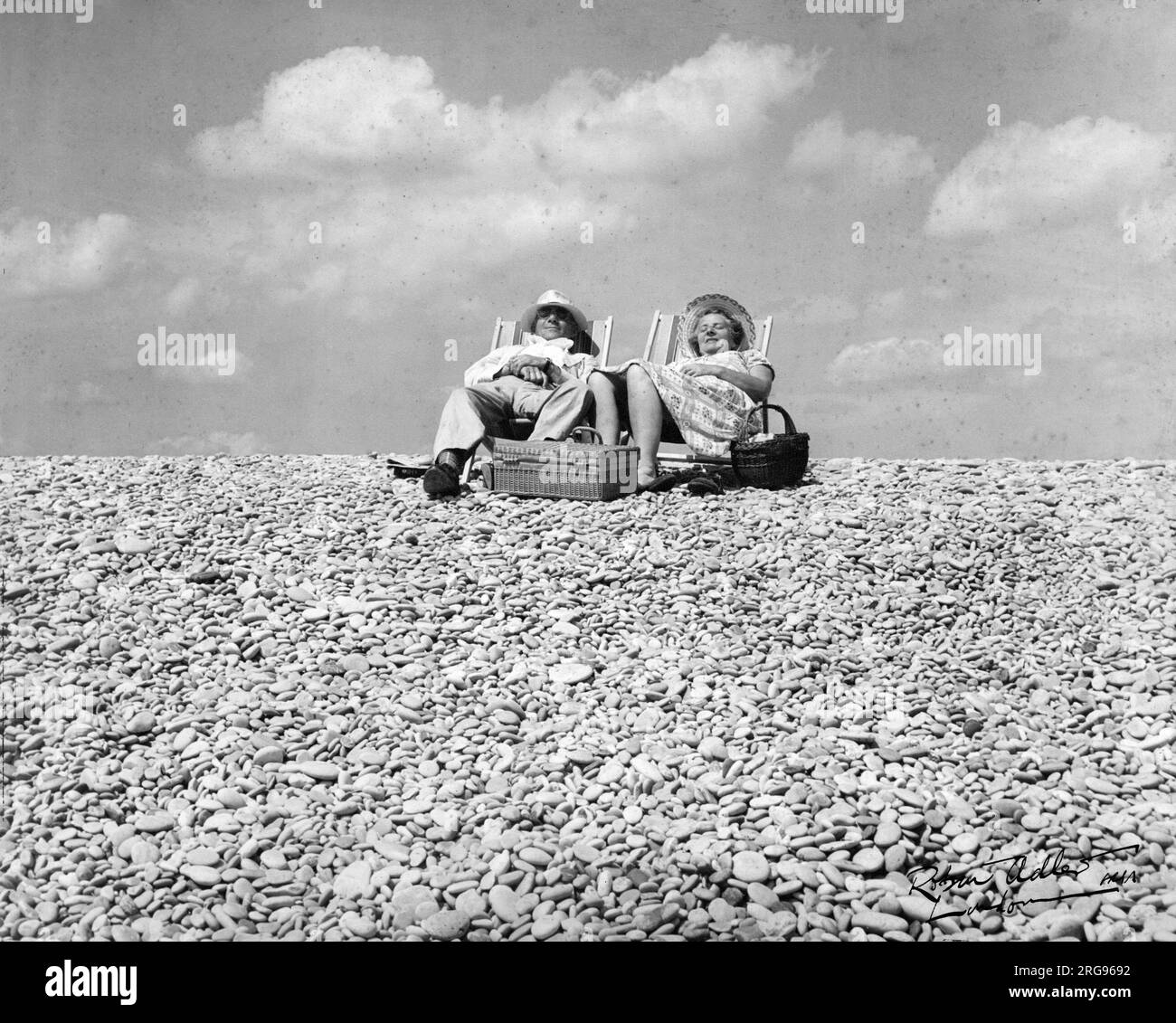 Ein zufriedenes Paar mittleren Alters am Meer, das auf Liegestühlen an einem Kieselstrand mit einem Picknickkorb sitzt. Stockfoto
