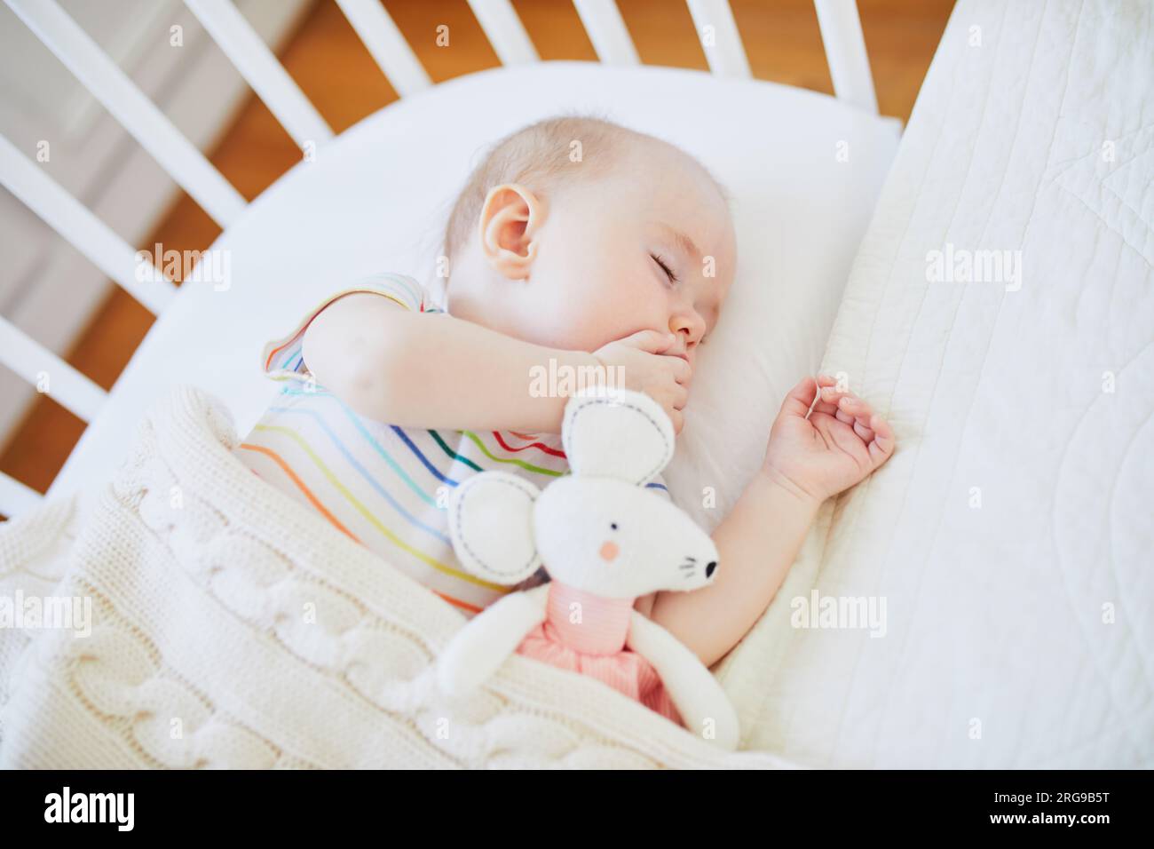 Ein bezauberndes Mädchen, das in einem Babybett schläft, das mit Plüschspielzeug am Bett der Eltern befestigt ist. Ein kleines Kind schläft einen Tag im Kinderbett. Das Schlaftrainingskonzept Stockfoto