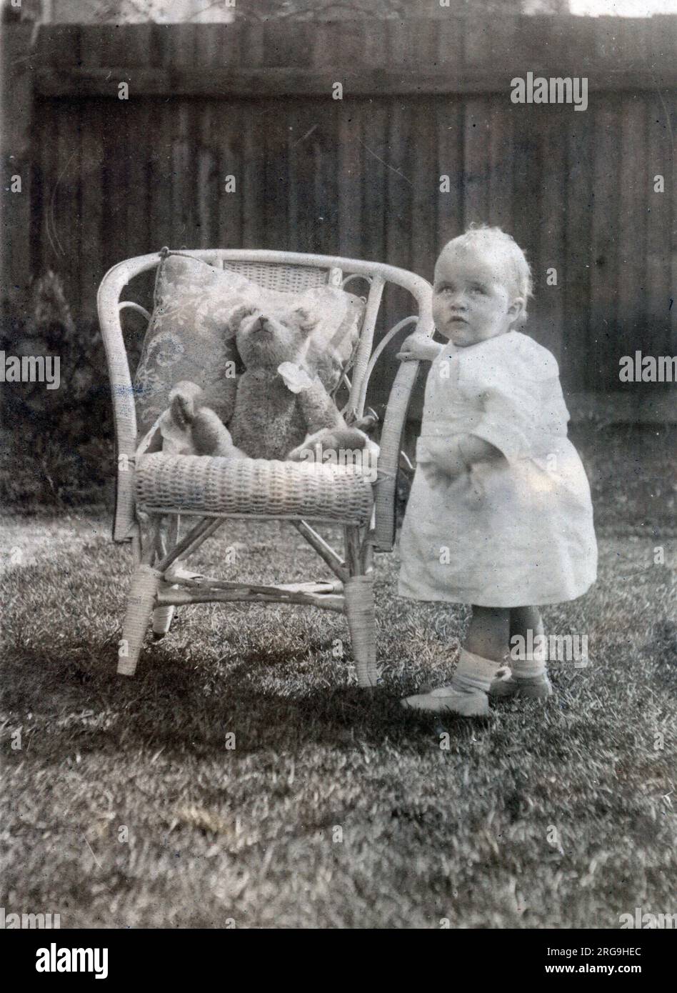 Ein Baby krabbelt in seinem Garten zu einem eleganten Rohrstuhl, um ihren Lieblings-Teddybären zu begrüßen. Stockfoto