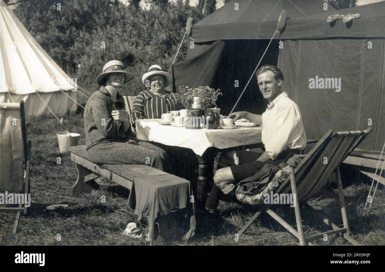 Bild 1/2 - Ein fröhliches Trio (ein junges Paar und die Mutter der jungen Dame?) Genießen Sie ein sehr schickes Frühstück auf dem sehr gut ausgestatteten Campingplatz, der komplett mit komplettem porzellanservice, elegantem Esstisch und schicken Zelten ausgestattet ist! Stockfoto