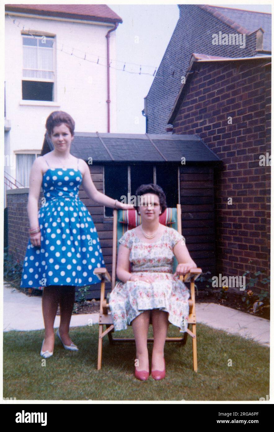 Eine Mutter (sitzend in einem Holz- und Leinwandklappstuhl) und ihre Tochter (längs stehend, in einem blau-weißen Polka-Dot-Kleid) posieren (eher styfly) für ein Foto in einem gepflegten britischen Vorstadtgarten - August 1960 Stockfoto