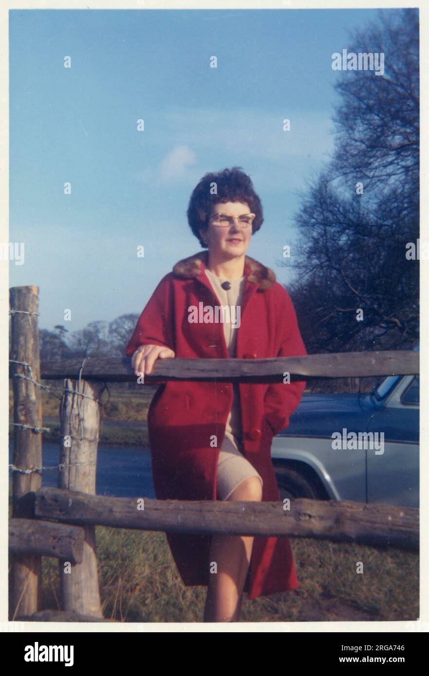 Eine Frau mittleren Alters in einem roten Mantel, die für ein Foto an einem Holzzaun posiert. Sie muss auf den Stacheldraht aufpassen...! April 1964 Stockfoto