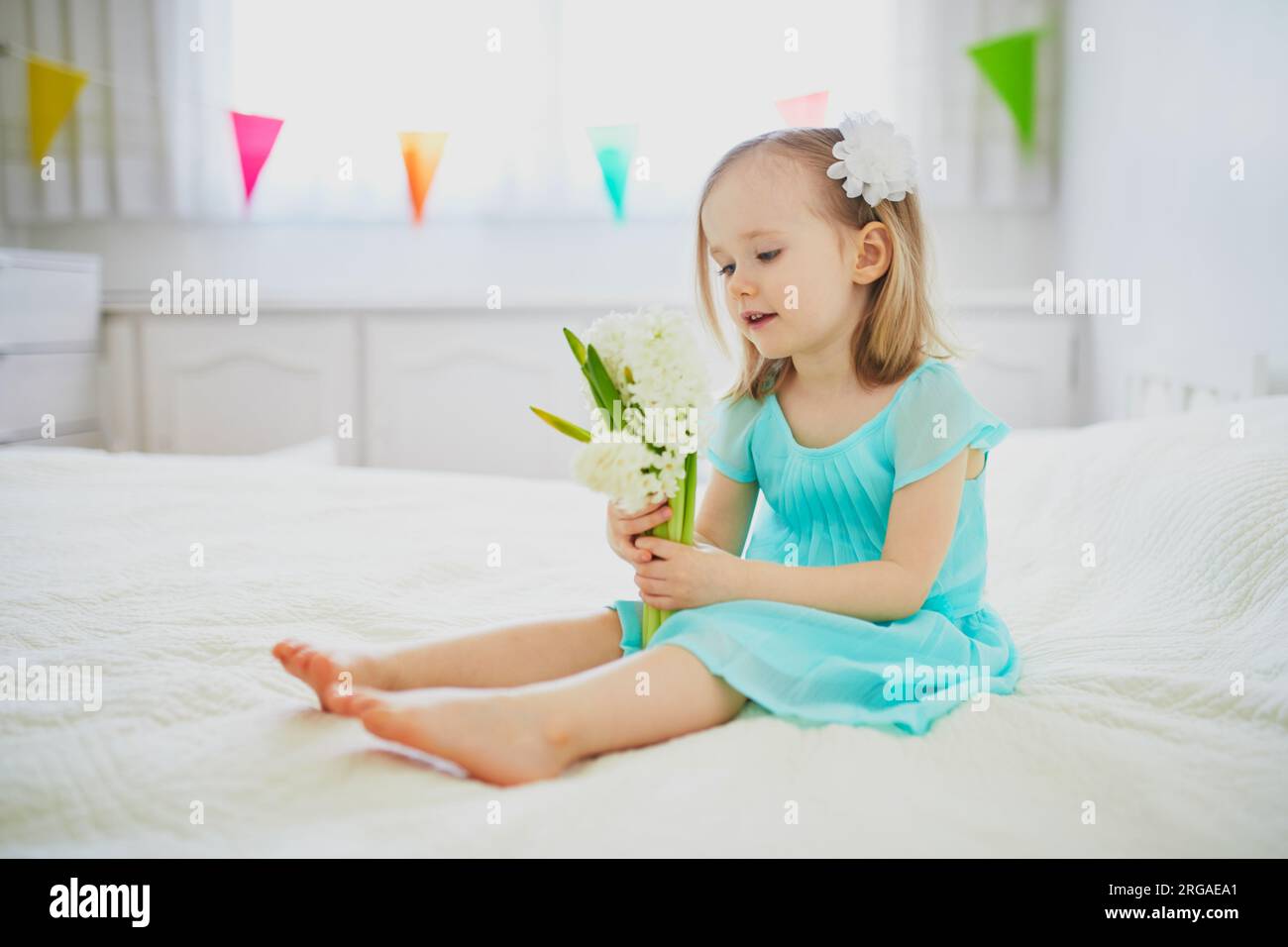 Ein bezauberndes Kleinkind, das mit weißen Hyazinthen auf dem Bett sitzt. Kleinkinder feiern Geburtstag Stockfoto