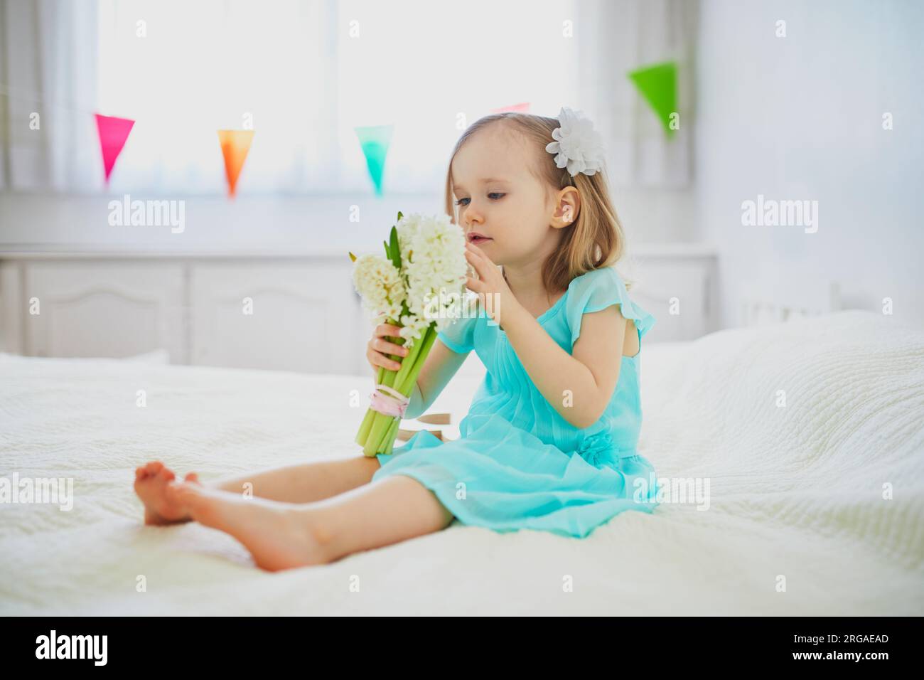 Ein bezauberndes Kleinkind, das mit weißen Hyazinthen auf dem Bett sitzt. Kleinkinder feiern Geburtstag Stockfoto