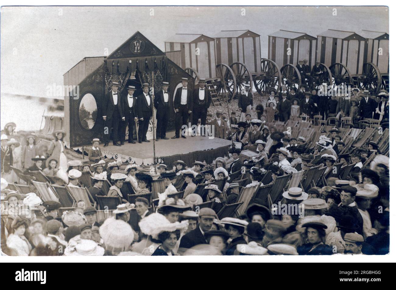 Dieses hervorragende Bild zeigt sechs Kostüme mit Strohhüten auf der Bühne vor einem Publikum mit Deck-Vorsitz. Behined The Stage sind fünf rollende Strandhütten in gestreifter Lackierung. Stockfoto