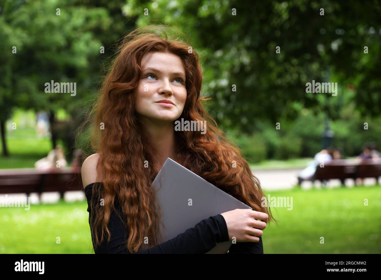 Lächelndes Mädchen mit langen roten Haaren und Sommersprossen, das im Sommerpark mit einem Laptop in der Hand stand. Lernen und Kreativität in der Natur Stockfoto