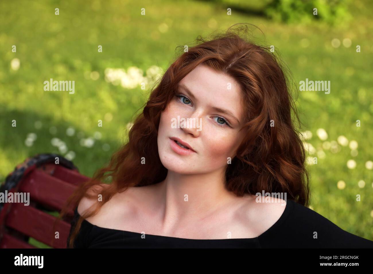 Attraktives Mädchen mit langen roten Haaren, das auf einer Bank im Sommerpark sitzt. Ein junges sinnliches Modell, weibliche Schönheit Stockfoto