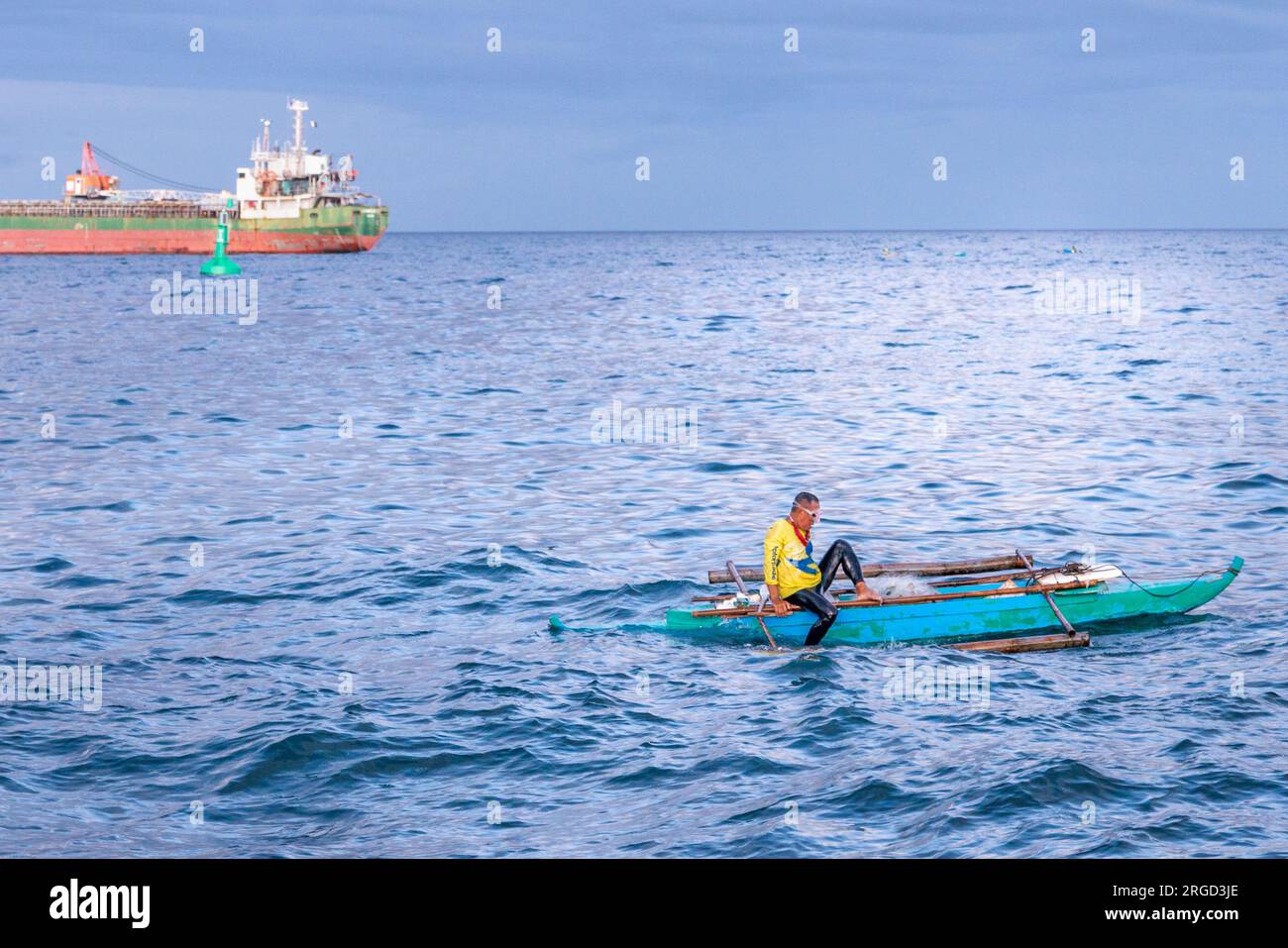 Dumaguete, Negros Island, Philippinen-Februar 01 2023: In der Dämmerung wirft der Mann Netze und taucht ins Wasser für Fische und ein großes Schiff ist nahe vor Anker Stockfoto