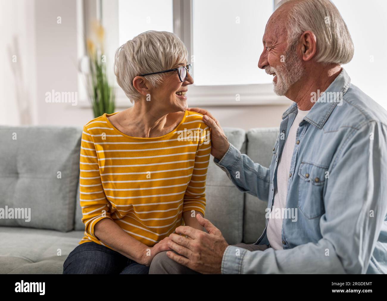 Seniorenpaar sitzt auf dem Sofa im Wohnzimmer und redet und Mann hält seine Hand an der Schulter einer Frau als Stützzeichen Stockfoto