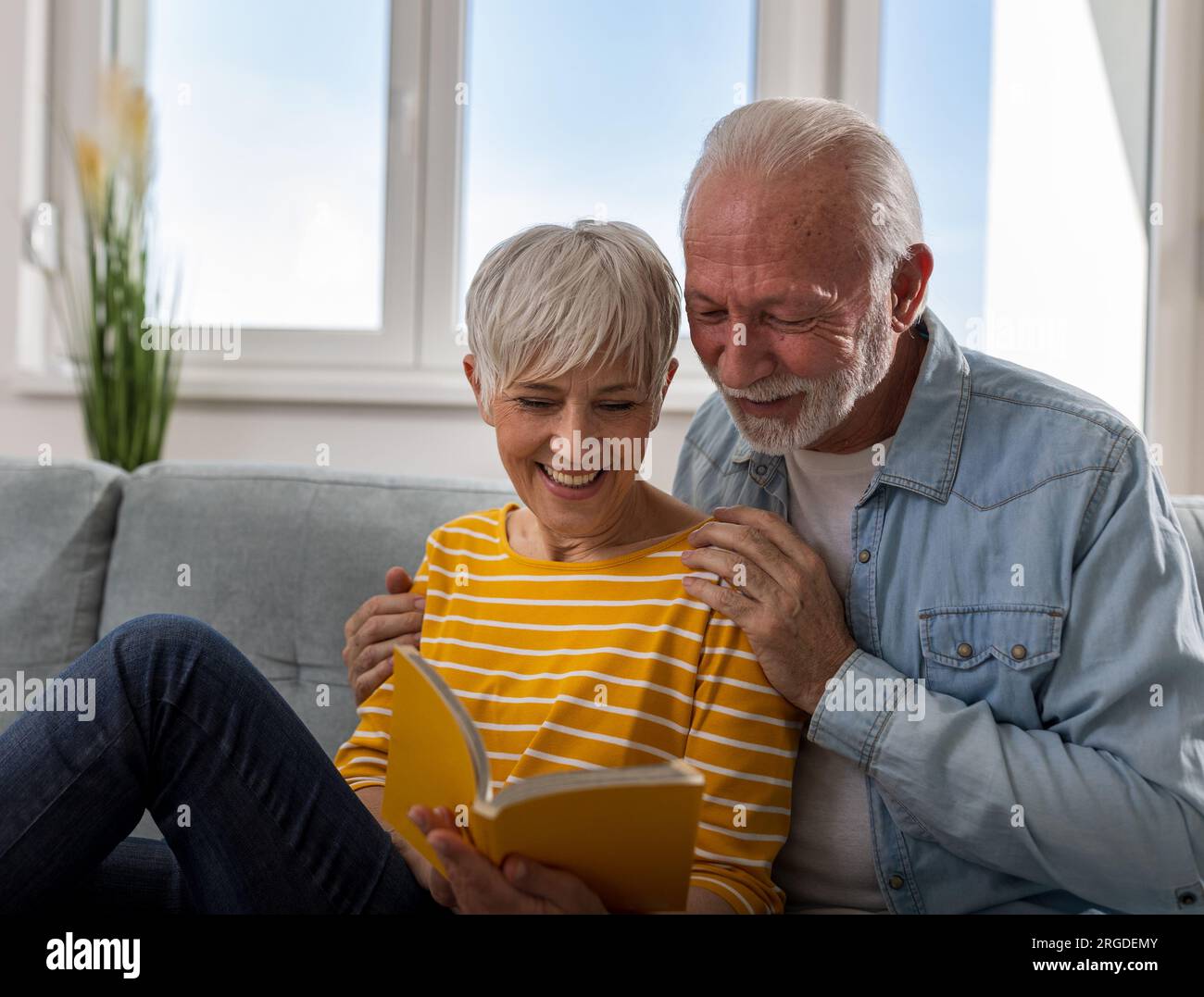 Fröhliches Seniorenpaar, das zu Hause auf dem Sofa sitzt, zusammen ein Buch liest und lächelt Stockfoto