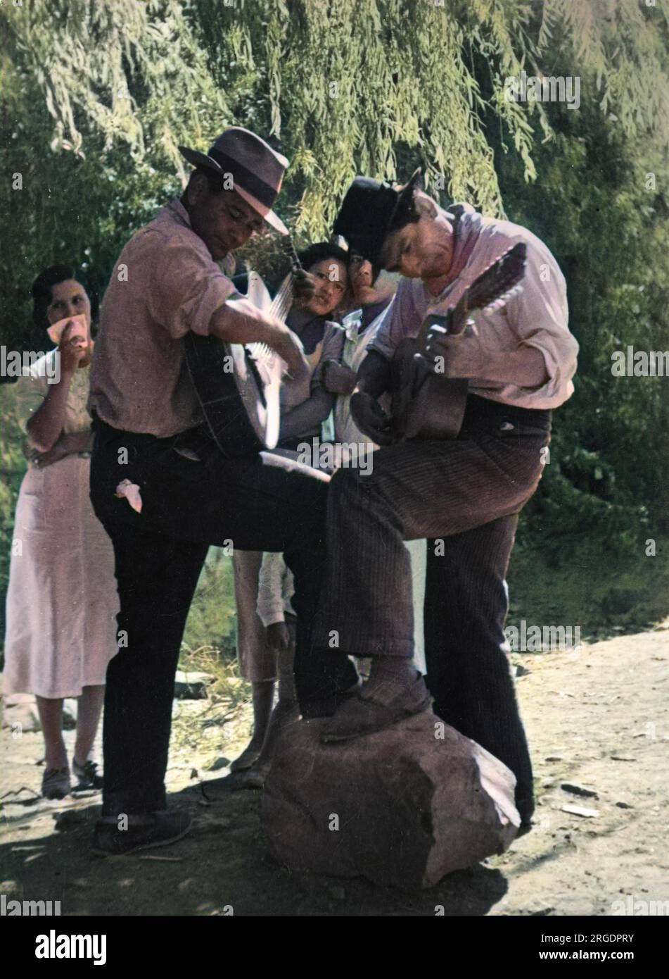 Argentinische Gitarrenspieler treten mit einem Fuß auf einen Stein in eine männliche Pose, während sie zusammen ein Duett spielen, während sich ihre schüchternen Frauen vor der Kamera, Südamerika, verstecken. Stockfoto