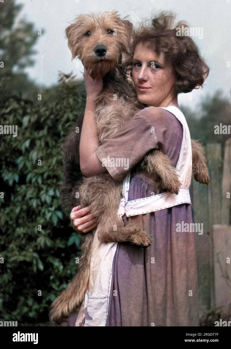 Eine schlanke junge Frau, die eine Schürze trägt und einen Hund in einem Garten hält. Stockfoto