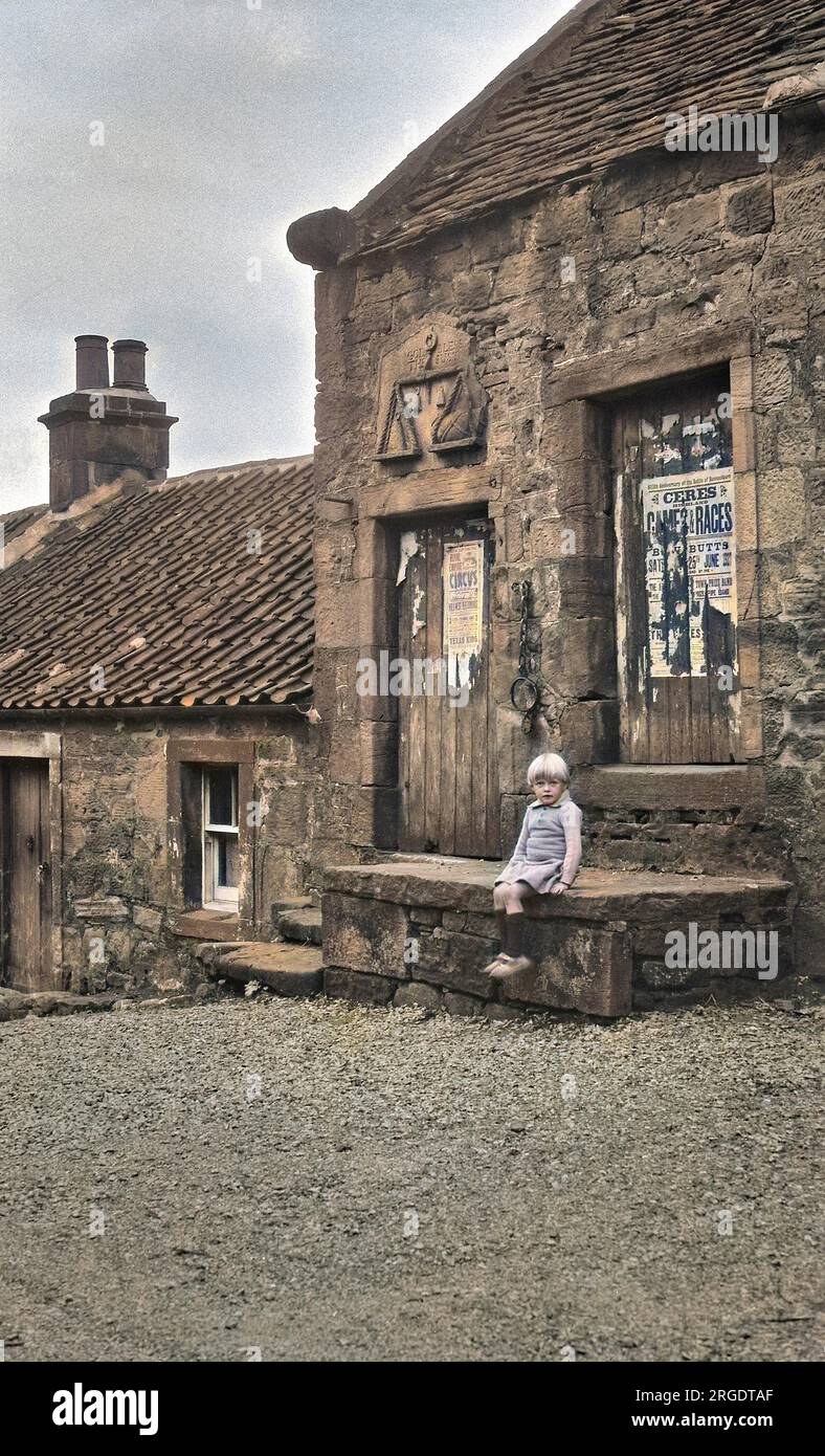 Straßenszene in den schottischen Highlands, mit einem kleinen Mädchen, das auf einer Steintreppe sitzt. Eine Steinschnitzerei einer Waage trägt die Inschrift: Gott segne die Gerechten. Stockfoto
