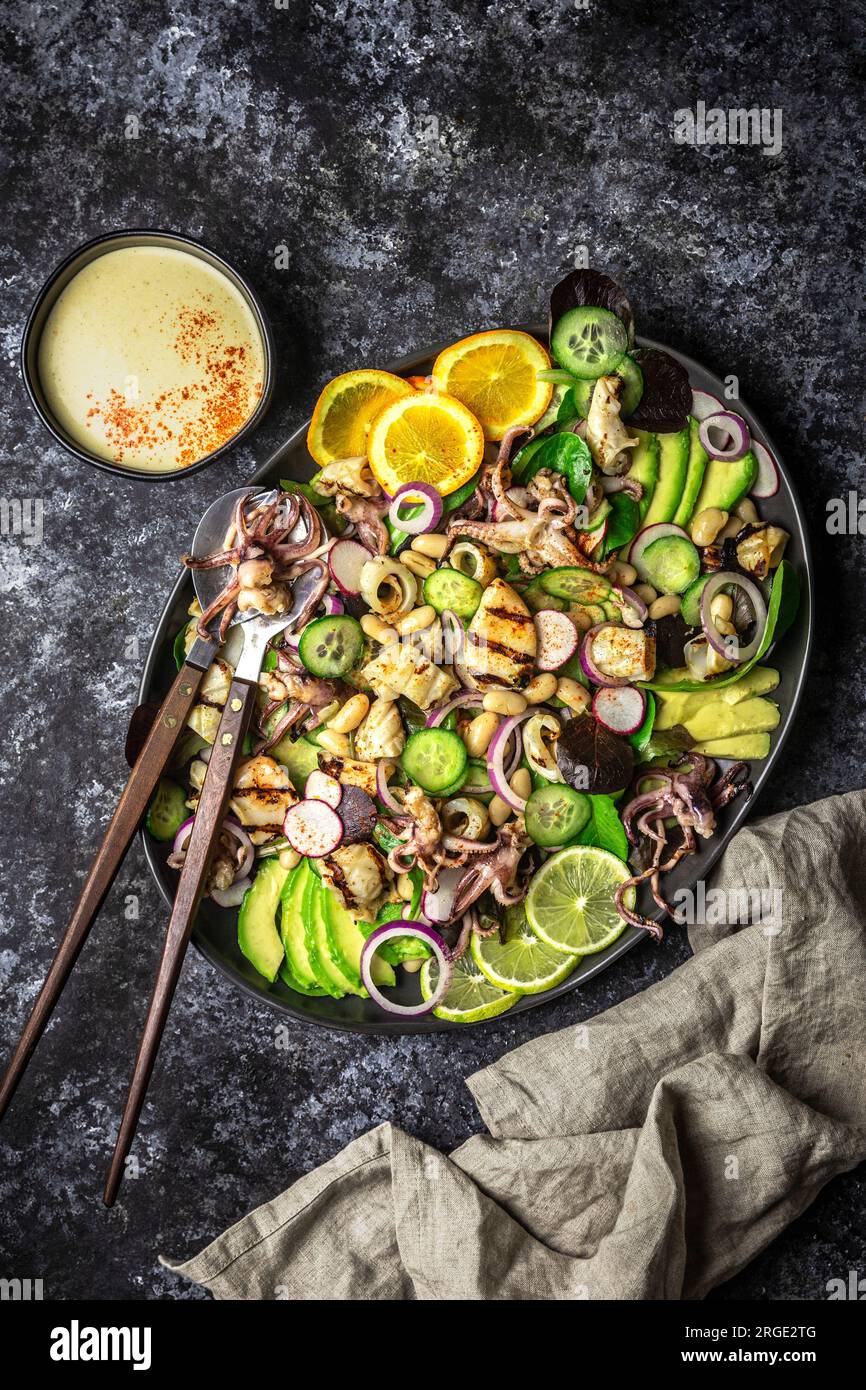 Gegrillter Tintenfischsalat mit Orange, Limette und Avocado auf einer dunklen Platte mit Sauce-Schüssel Stockfoto