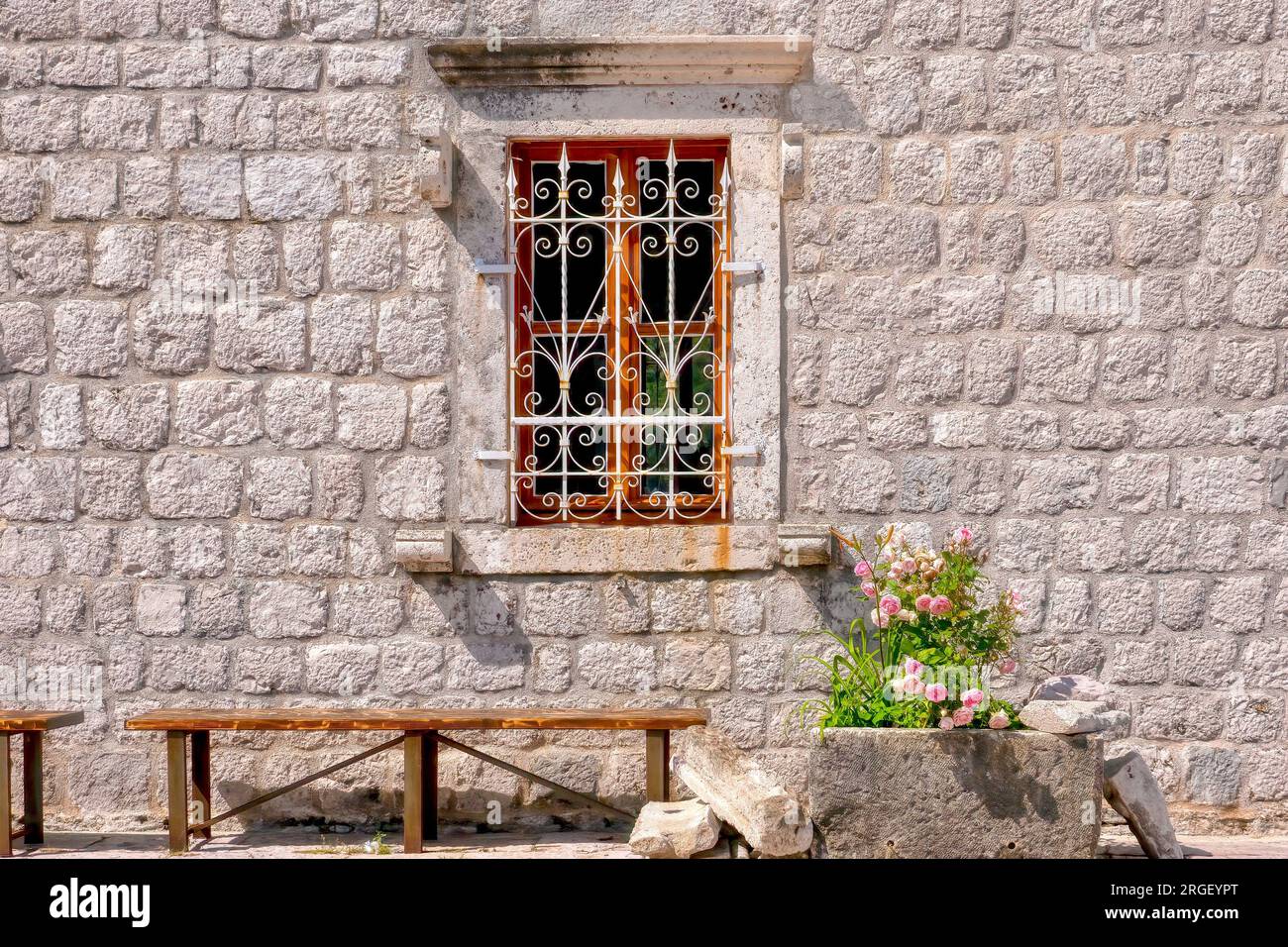 Ein malerisches Fenster mit kunstvoll verziertem schmiedeeisernem Gitter in einer alten Steinwand, mit einer Holzbank und pinkfarbenen Rosen davor. In Montenegro. Stockfoto