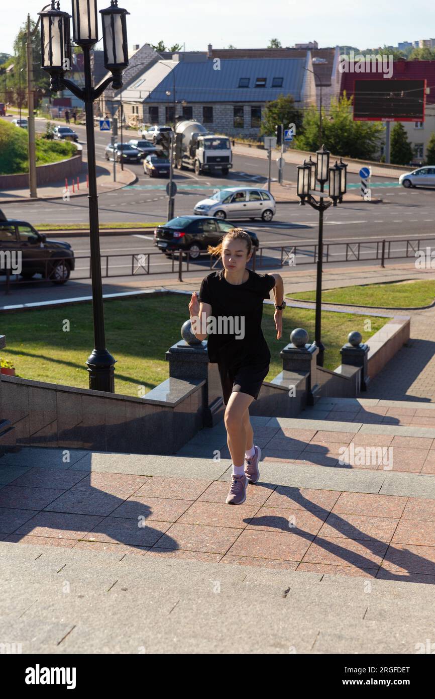 Eine junge Frau in schwarzen Klamotten, die früh morgens auf der Stadtstraße auf der Treppe rennt. Stockfoto