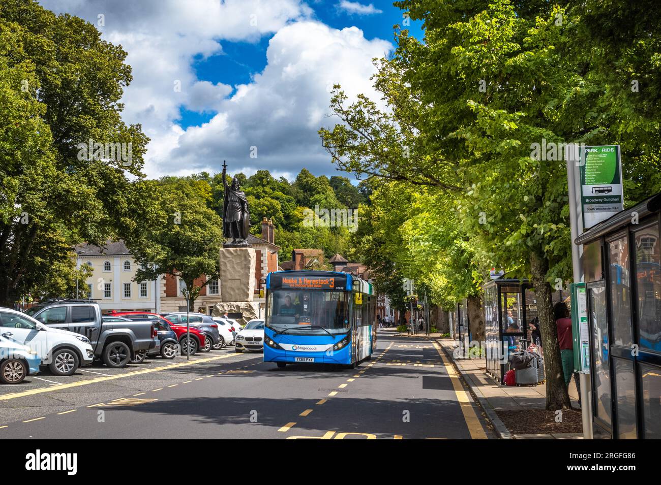 Ein blauer Eindeckbus fährt an Bushaltestellen vorbei, neben der riesigen Statue von König Alfred dem Großen am Broadway in Winchester, Hampshire, Großbritannien. Stockfoto