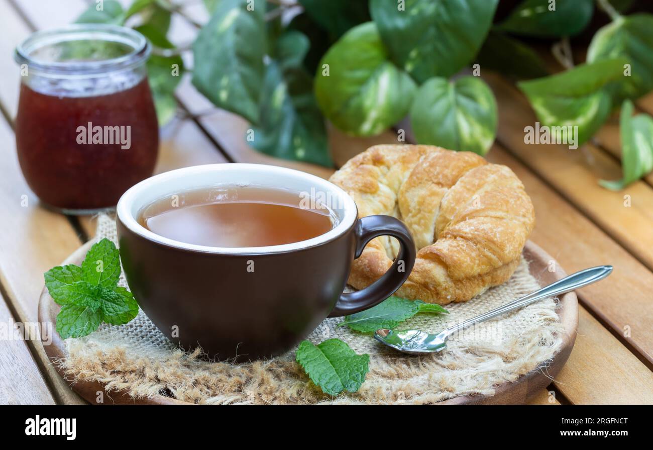 Tasse Tee mit Minzeblättern, Croissant und Marmelade draußen auf einem hölzernen Terrassentisch Stockfoto