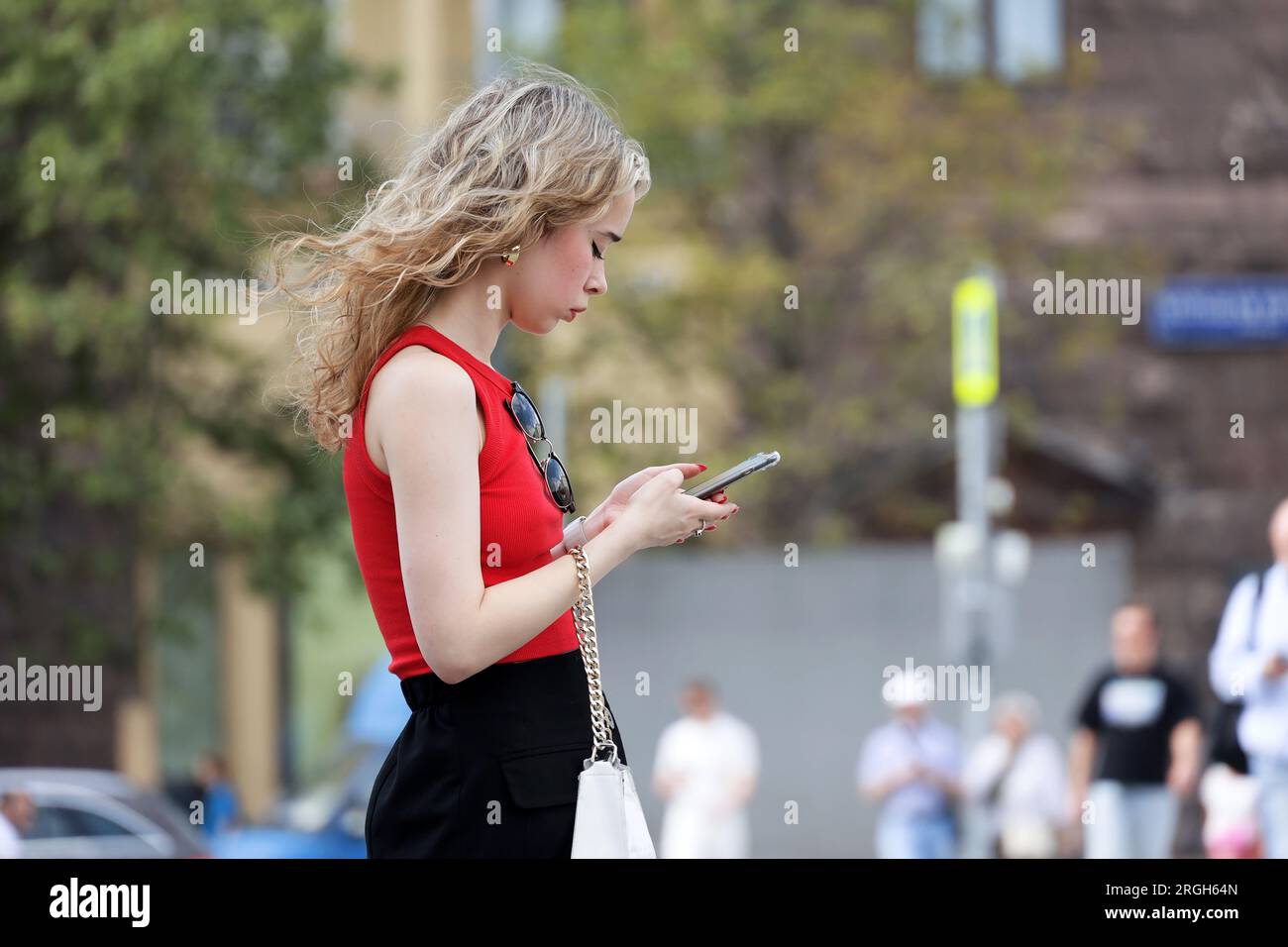 Attraktives Mädchen mit blonden Locken, das auf einer Straße mit einem Smartphone in der Hand steht und in der Sommerstadt ein Mobiltelefon benutzt Stockfoto