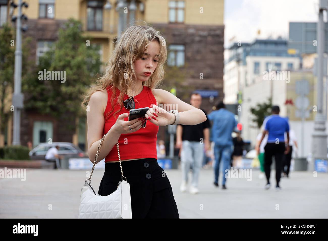Attraktives Mädchen mit blonden Locken, das auf einer Straße mit einem Smartphone in der Hand steht und in der Sommerstadt ein Mobiltelefon benutzt Stockfoto