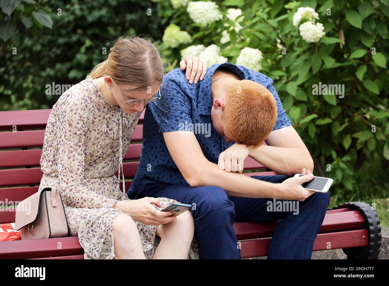 Ein Paar sitzt mit Smartphones auf einer Holzbank im Sommerpark, ein rothaariger Mann und ein Mädchen mit Brille Stockfoto