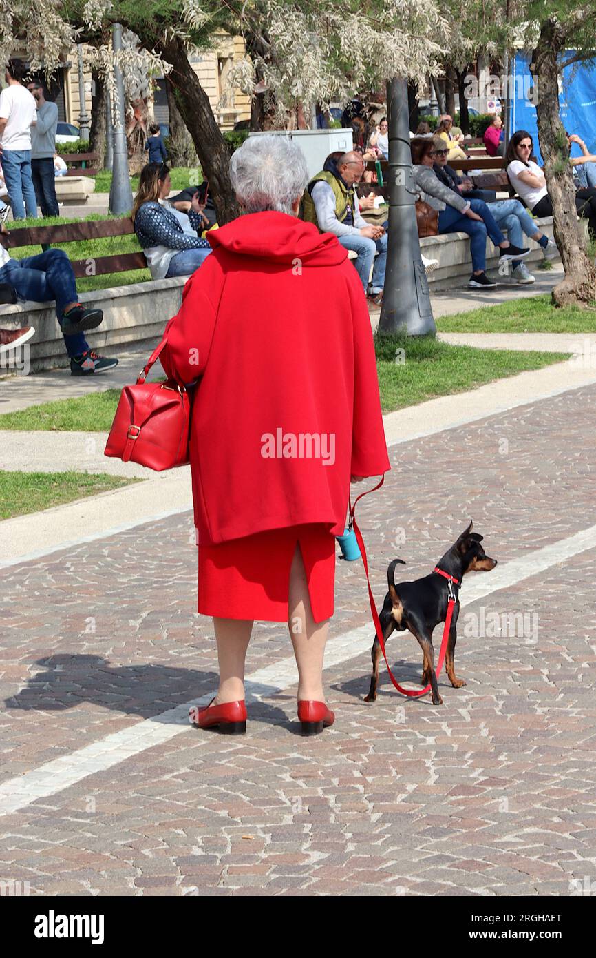 Eine elegant gekleidete ältere Dame in scharlachrot mit passenden Accessoires führt ihren Hund in hellem Sonnenschein am Meer in Salerno, Italien entlang. Stockfoto