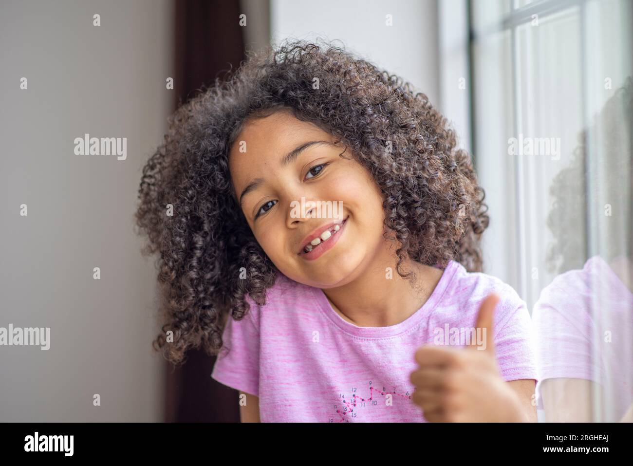 Wunderschönes kleines glückliches Mädchen, das neben dem Fenster sitzt, Nahaufnahme eines fröhlichen, lockigen Kindes, das den Daumen zeigt Stockfoto