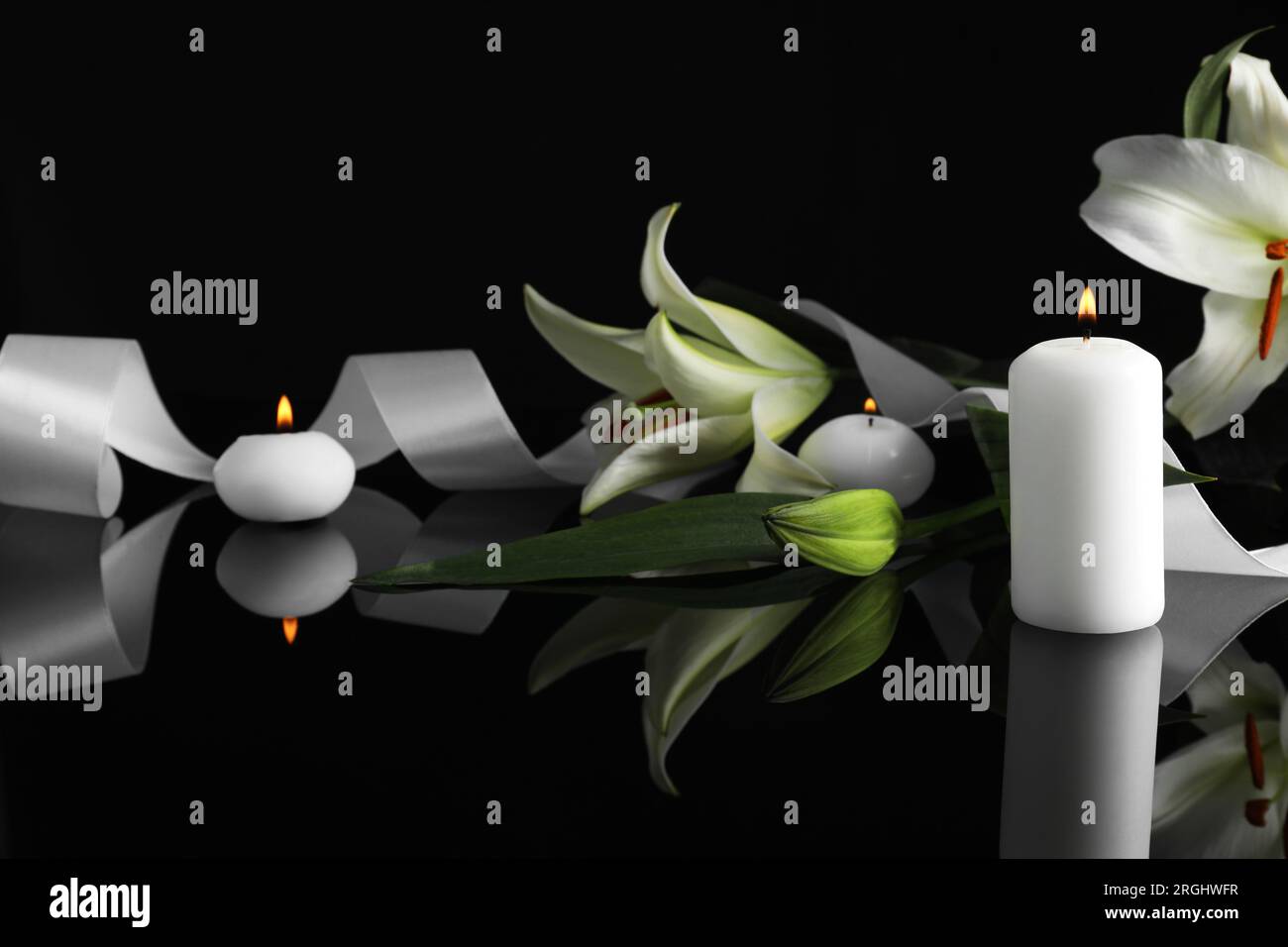 Weiße Lilien und brennende Kerzen auf schwarzer Spiegeloberfläche bei Dunkelheit, Platz für Text. Begräbnissymbole Stockfoto