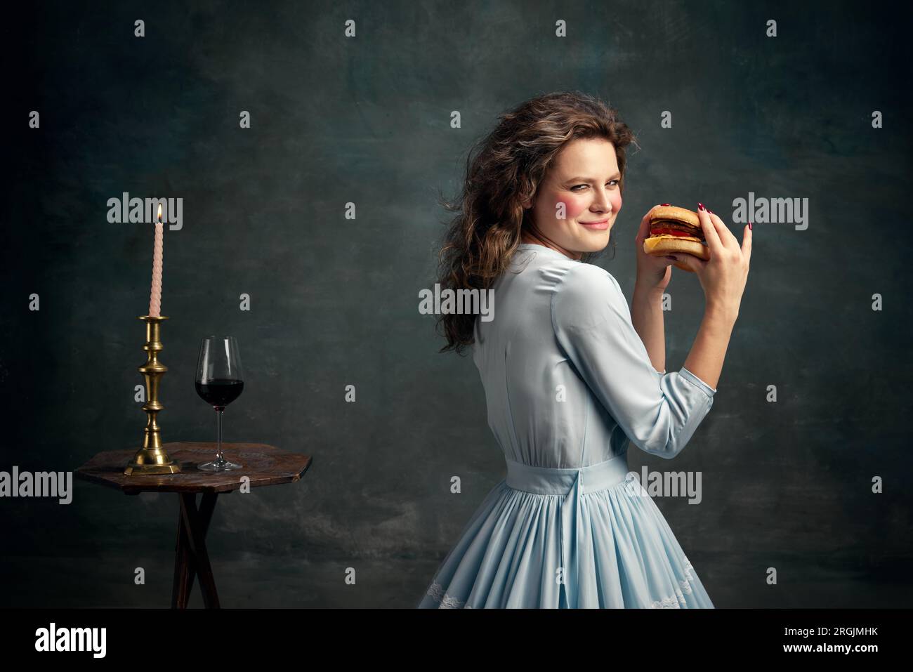 Prinzessin. Porträt einer aristokratischen schlau aussehenden Frau, die ein blaues historisches Kleid trägt und einen heißen, großen Hamburger hält. Moderne Esskunst. Stockfoto