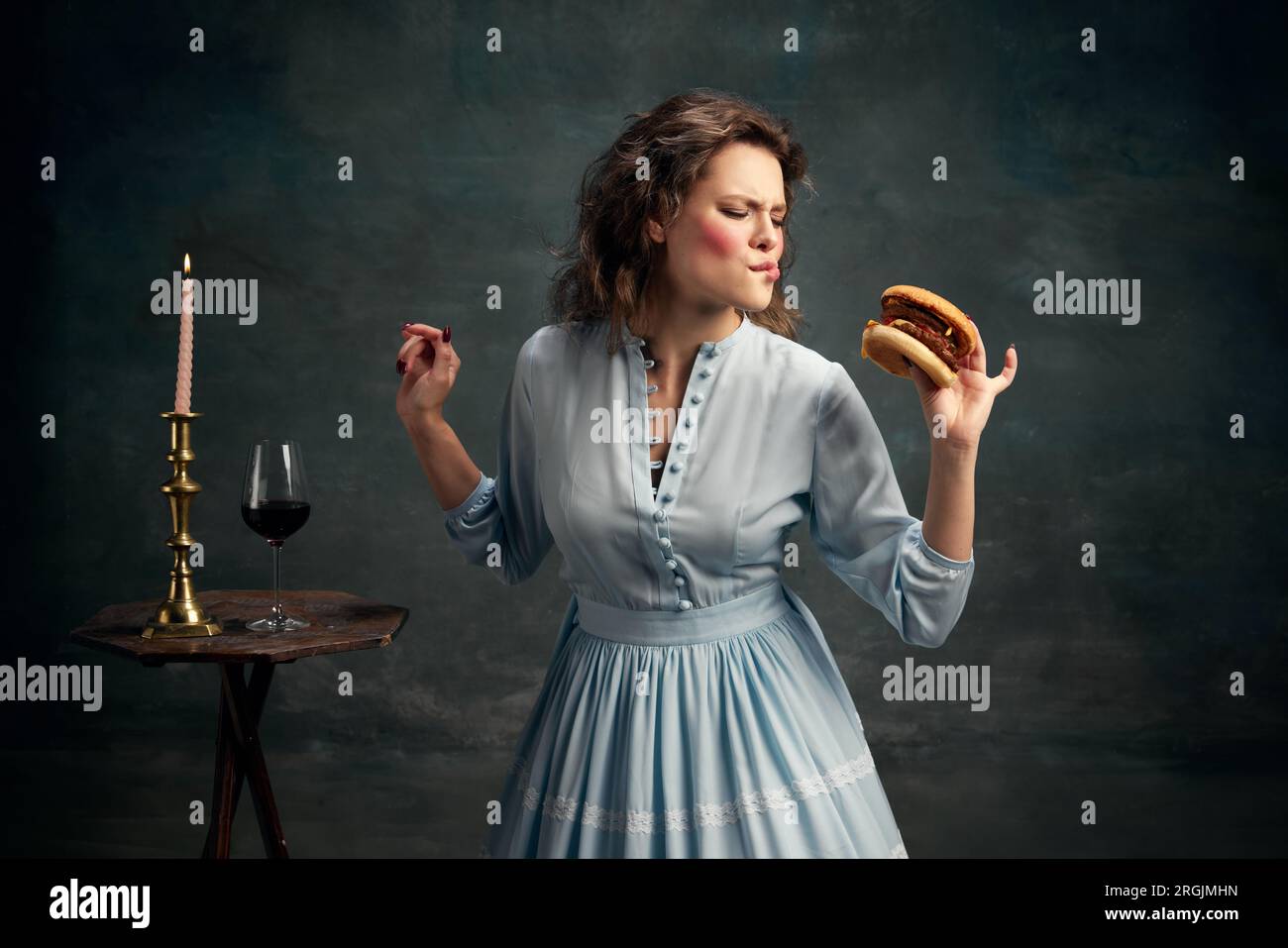 Prinzessin. Porträt einer aristokratischen hungrigen Frau, die ein blaues historisches Kleid trägt und großen Hamburger isst. Moderne Esskunst. Stockfoto