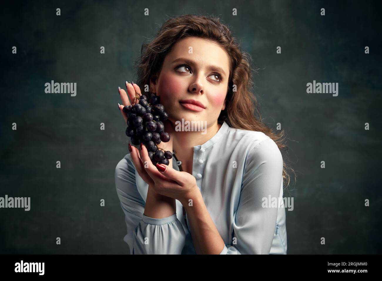 Wunderschöne, glamouröse junge Frau in einem blauen historischen Kleid, die Trauben präsentiert und wegschaut. Essensstimmung. ERAS-Vergleich Stockfoto