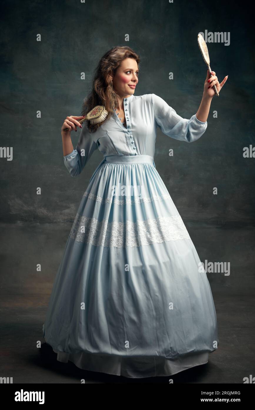 Ein vollständiges Porträt einer charmanten Aristokratin, die ein blaues historisches Kleid trägt, einen alten Spiegel in der Hand hält und Haare bürstet. Stockfoto